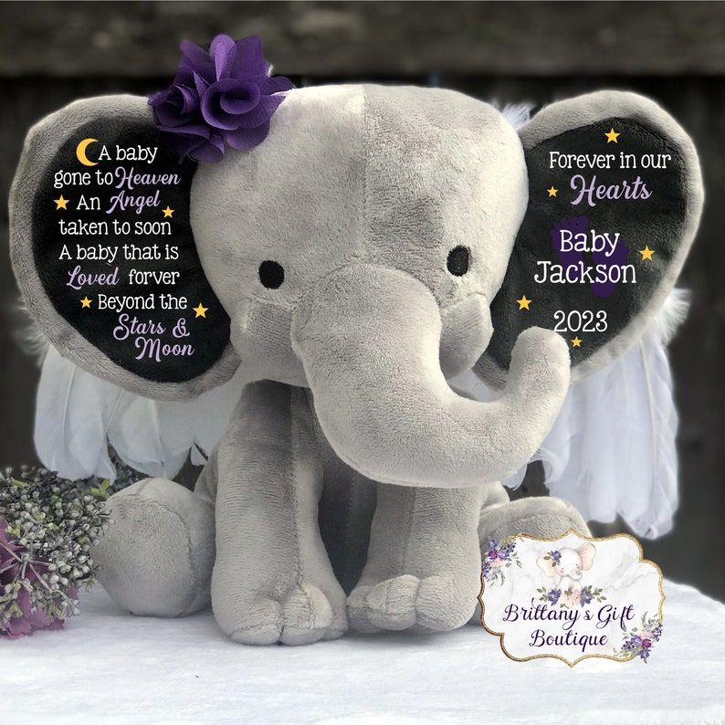 Puede incluir: Un elefante de peluche gris con alas, una flor morada en la cabeza y parches negros en las orejas. Los parches tienen texto que dice "A baby gone to Heaven, An Angel taken to soon, A baby that is Loved forever, Beyond the Stars & Moon" y "Forever in our Hearts, Baby Jackson, 2023".