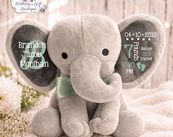 May include: A plush gray elephant toy featuring a sage green satin bow. The left ear displays the name Brandon Jacob Monihan with a large letter B. The right ear shows birth details: 04-10-2019, 7 Pounds 7 Ounces, 19.5 Inches, and 6:30 PM.