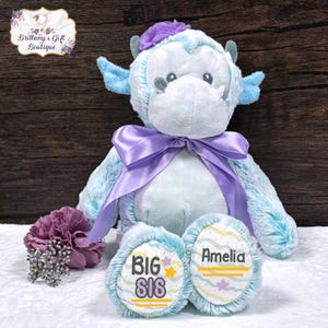 May include: A plush, light blue stuffed animal with a purple bow and flower accents. The bear has the words "BIG SIS" and the name "Amelia" printed on its feet. The background is a dark wood.