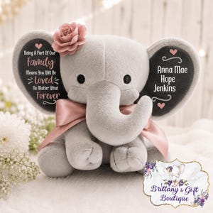 May include: A plush gray elephant toy featuring a pink satin ribbon around its neck and a pink fabric flower on its head. The left ear displays the text "Being A Part Of Our Family Means You Will Be Loved No Matter What Forever" and the right ear displays "Anna Mae Hope Jenkins".