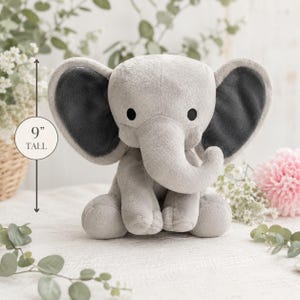 Puede incluir: Un peluche de elefante gris, de 23 cm de altura, con orejas grandes y una trompa. El elefante tiene ojos negros y est&aacute; sentado sobre una superficie blanca. El fondo incluye vegetaci&oacute;n y una flor rosa.