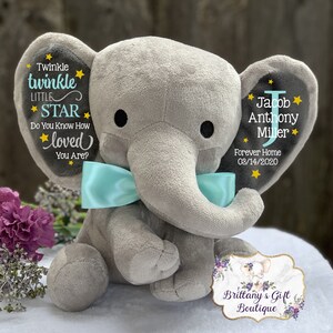 May include: A plush gray elephant toy featuring a light blue satin bow around its neck. The left ear displays the text Twinkle twinkle LITTLE STAR Do You Know How loved You Are? with yellow stars. The right ear shows the name Jacob Anthony Miller.