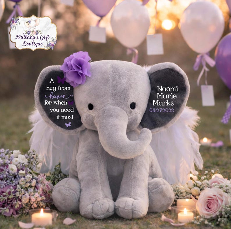 Puede incluir: Un elefante de peluche gris con una flor morada en la cabeza y alas de plumas blancas. El elefante tiene texto en las orejas que dice "A hug from heaven for when you need it most" y "Naomi Marie Marks 03/27/2022". Rodeado de flores y velas.