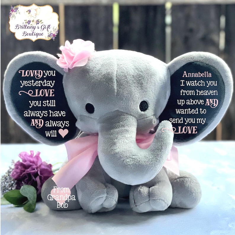 Puede incluir: Un elefante de peluche gris con orejas rosas y un lazo rosa. El elefante tiene mensajes bordados en sus orejas que dicen "Loved you yesterday Love you still always have and always will" y "Annabella I watch you from heaven up above and wanted to send you my Love". El elefante tambi&eacute;n tiene una flor rosa en la cabeza y las palabras "From Grandpa Bob" en su pie.