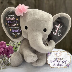 Puede incluir: Un elefante de peluche gris con una flor rosa en la cabeza. Las orejas del elefante tienen el texto "Twinkle Twinkle Little Star" y un nombre. El elefante est&aacute; sentado sobre una superficie blanca.