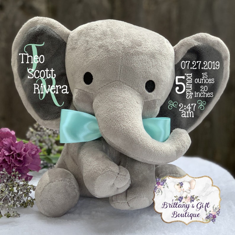 May include: A gray plush elephant with a light blue bow, personalized with the name "Theo Scott Rivera" and birth details: 5 pounds, 15 ounces, 20 inches, and the date and time of birth. The elephant has black eyes.