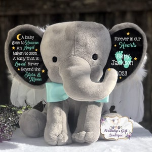 Puede incluir: Un elefante de peluche gris con alas y un lazo turquesa. El elefante tiene un parche de fieltro negro en su oreja con el texto "A baby gone to Heaven ★ An Angel taken to soon A baby that is Loved forever ★ Beyond the Stars & Moon". La otra oreja tiene un parche de fieltro negro con el texto "Forever in our ★ Hearts Baby Jackson ★ 2023".