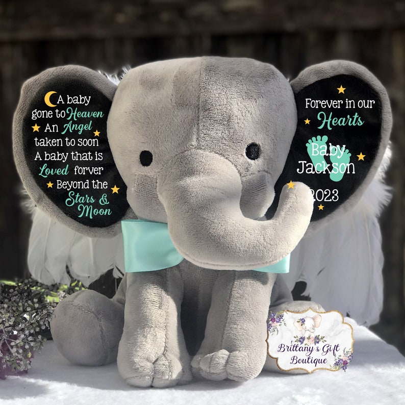 Puede incluir: Un elefante de peluche gris con alas y un lazo turquesa. El elefante tiene un parche de fieltro negro en su oreja con el texto "A baby gone to Heaven ★ An Angel taken to soon A baby that is Loved forever ★ Beyond the Stars & Moon". La otra oreja tiene un parche de fieltro negro con el texto "Forever in our ★ Hearts Baby Jackson ★ 2023".
