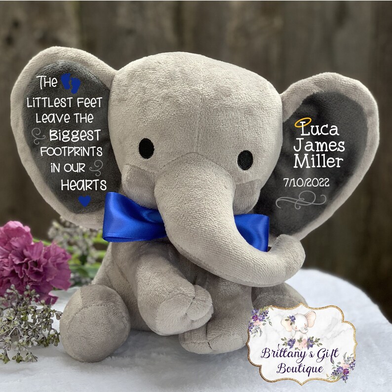 Op de afbeelding: Een grijze pluche olifant met een blauwe strik. De oren van de olifant zijn geborduurd met de tekst "The littlest feet leave the biggest footprints in our hearts" en "Luca James Miller 7/10/2022".