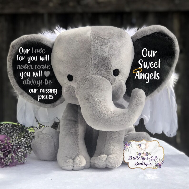 Może przedstawiać: Szary pluszowy słoń z białymi skrzydłami i czarnymi uszami. Jedno ucho ma napis "Our Love for you will never cease you will always be our missing pieces" z sercem. Drugie ucho ma napis "Our Sweet Angels".