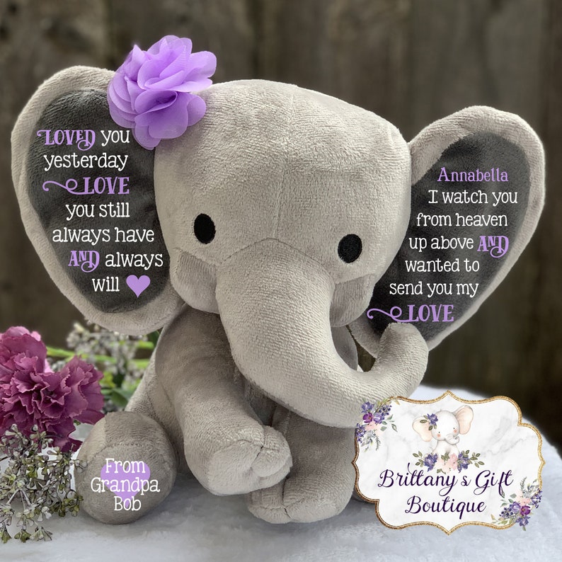 Puede incluir: Un elefante de peluche gris con detalles de flores moradas. El elefante tiene texto bordado en ambas orejas, que dice "Loved you yesterday, Love you still always have and always will" y "Annabella, I watch you from heaven up above and wanted to send you my Love". El elefante est&aacute; sentado sobre una superficie blanca con un dise&ntilde;o floral y el texto "From Grandpa Bob" en un lado y "Brittany's Gift Boutique" en el otro.