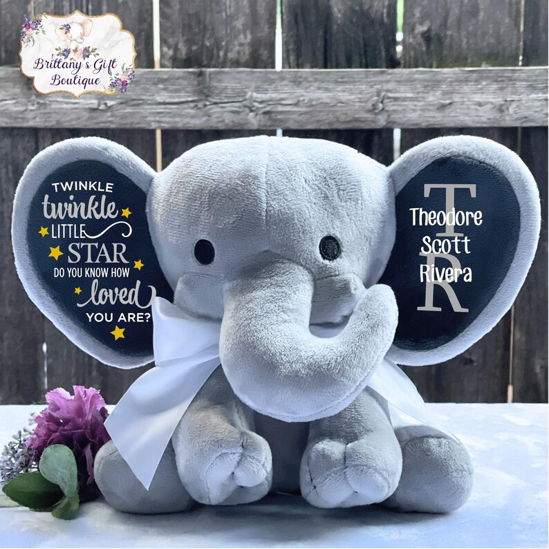 Puede incluir: Un elefante de peluche gris con un lazo blanco. Las orejas del elefante tienen texto personalizado: una dice "Twinkle Twinkle Little Star, do you know how loved you are?" y la otra muestra el nombre "Theodore Scott Rivera".