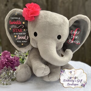 Puede incluir: Un peluche de elefante gris con ojos negros y una flor rosa en la cabeza. Las orejas del elefante tienen texto, incluyendo "Twinkle Twinkle Little Star" y un nombre. Tambi&eacute;n es visible un logotipo de una boutique de regalos.