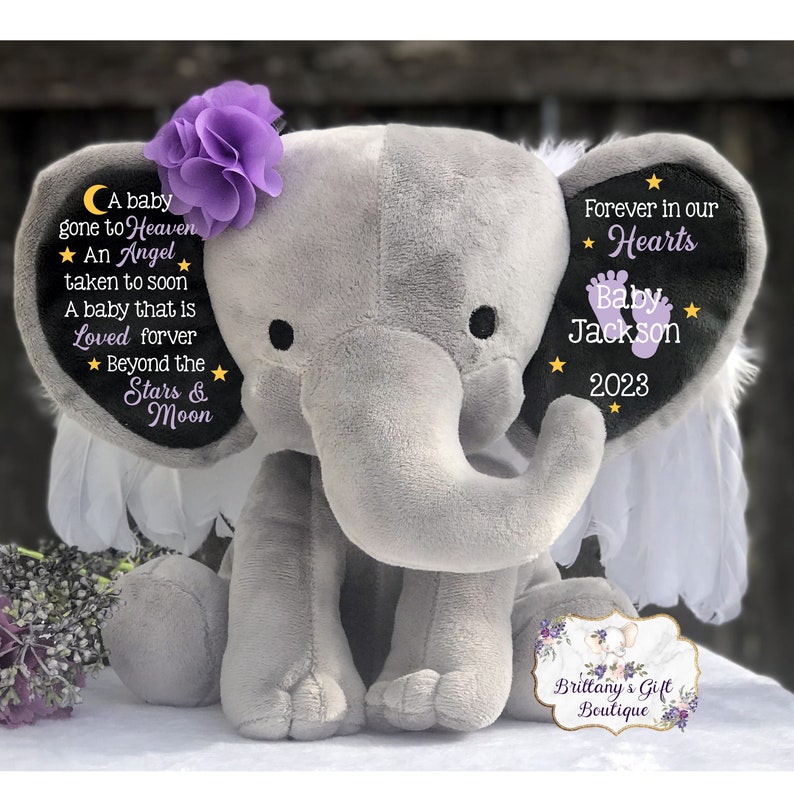 Puede incluir: Un elefante de peluche gris con alas moradas y una flor morada en la cabeza. El elefante tiene un parche negro en la oreja con el texto "Forever in our Hearts Baby Jackson 2023".
