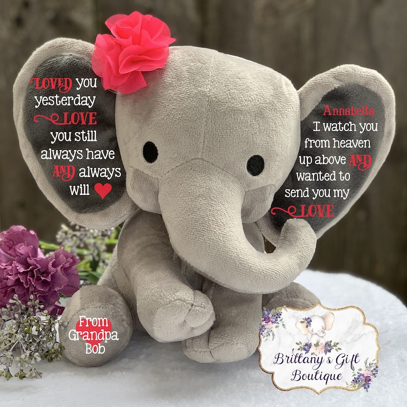 Puede incluir: Un elefante de peluche gris con una flor rosa en la cabeza. El elefante tiene texto bordado en sus orejas que dice "Loved you yesterday, Love you still, always have and always will" y "Annabella, I watch you from heaven up above and wanted to send you my love". El elefante est&aacute; sentado en una superficie blanca con una peque&ntilde;a etiqueta redonda que dice "From Grandpa Bob".