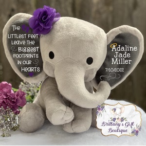 Op de afbeelding: Een grijze pluche olifant met paarse bloemenaccenten. Het oor van de olifant heeft de tekst "The littlest feet leave the biggest footprints in our hearts" en het andere oor heeft de tekst "Adaline Jade Miller 7/10/2022".