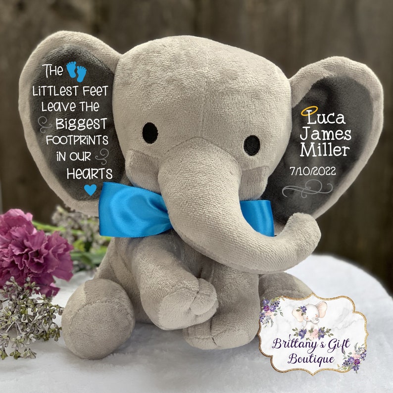 Op de afbeelding: Een grijze pluche olifant met een blauwe strik. Het oor van de olifant heeft de tekst "Luca James Miller 7/10/2022" geborduurd. Het andere oor heeft de tekst "The littlest feet leave the biggest footprints in our hearts" geborduurd.