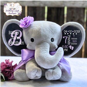 May include: A gray plush elephant toy featuring a purple satin ribbon around its neck and a purple flower on its head. The left ear displays the name Bailey Jane Monihan, while the right ear shows birth details: 04-10-2019, 7 Pounds 7 Ounces, 19.5 Inches, 6:30 PM.