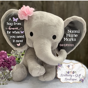 Puede incluir: Un elefante de peluche gris con orejas grandes, una trompa y ojos negros. La oreja izquierda tiene el texto "A hug from heaven for when you need it most" y gr&aacute;ficos de mariposas. La oreja derecha tiene el nombre "Naomi Marie Marks" y la fecha "03/27/2022". Una flor rosa adorna su cabeza.