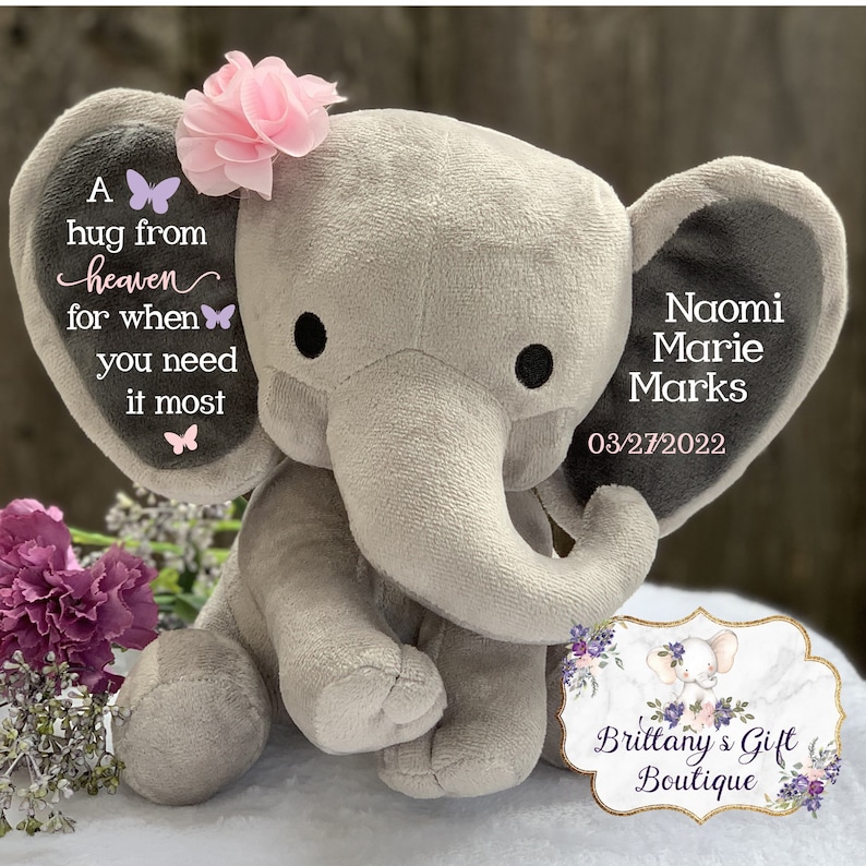 Puede incluir: Un elefante de peluche gris con orejas grandes, una trompa y ojos negros. La oreja izquierda tiene el texto "A hug from heaven for when you need it most" y gr&aacute;ficos de mariposas. La oreja derecha tiene el nombre "Naomi Marie Marks" y la fecha "03/27/2022". Una flor rosa adorna su cabeza.