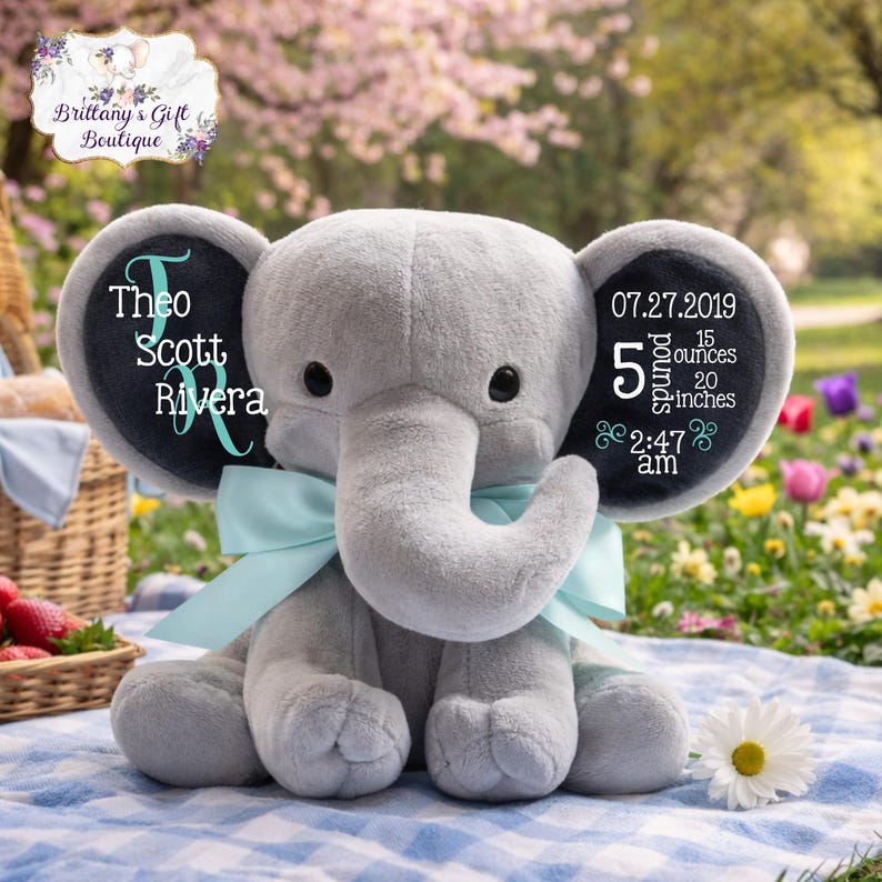 Puede incluir: Un elefante de peluche gris con un lazo azul claro. Las orejas del elefante tienen el texto "Theo Scott Rivera" y "07.27.2019, 2,27 kg, 50.8 cm, 2:47 am". El elefante est&aacute; sentado sobre una manta a cuadros azul y blanco.