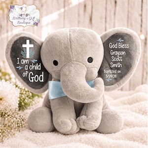 May include: A plush gray elephant toy with a light blue satin bow around its neck. The left ear features a white cross and the text I am a child of God. The right ear displays the text God Bless Grayson Scott Smith Baptized on 5/24/26.