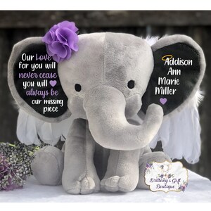 Może przedstawiać: Szary pluszowy słoń z skrzydłami i fioletowym kwiatem na głowie. Słoń ma czarne ucho z tekstem "Our Love for you will never cease you will always be our missing piece" i drugie czarne ucho z tekstem "Addison Ann Marie Miller".