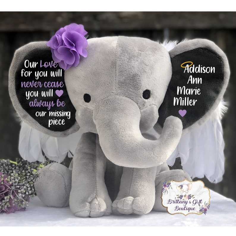 Może przedstawiać: Szary pluszowy słoń z skrzydłami i fioletowym kwiatem na głowie. Słoń ma czarne ucho z tekstem "Our Love for you will never cease you will always be our missing piece" i drugie czarne ucho z tekstem "Addison Ann Marie Miller".
