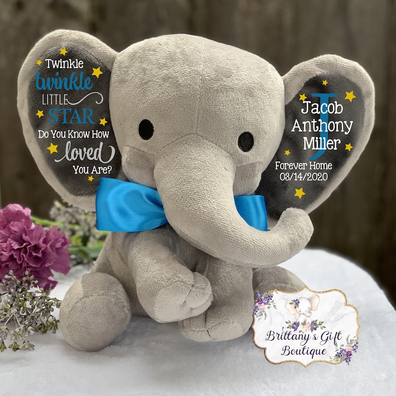 May include: A plush gray elephant toy with a bright blue satin bow around its neck. The left ear features the text Twinkle Twinkle Little Star Do You Know How Loved You Are? with yellow stars. The right ear displays the name Jacob Anthony Miller.