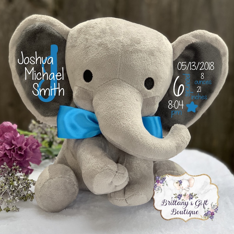 K&ouml;nnte beinhalten: Ein graues Pl&uuml;schelefant-Spielzeug mit einer leuchtend blauen Satinschleife um den Hals. Die Ohren sind mit Text bedruckt, darunter der Name Joshua Michael Smith, das Geburtsdatum 13.05.2018, die Uhrzeit 20:04 Uhr und Geburtsdaten von 6 Pfund, 8 Unzen.