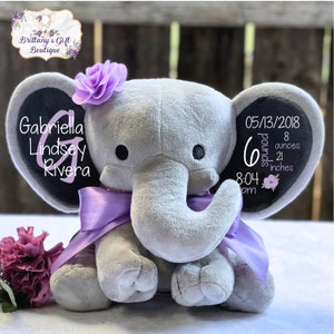 K&ouml;nnte beinhalten: Ein graues Pl&uuml;schelefant-Spielzeug mit einem violetten Satinband um den Hals und einer violetten Stoffblume auf dem Kopf. Das linke Ohr zeigt den Namen Gabriella Lindsey Rivera mit einem gro&szlig;en Buchstaben G, das rechte Ohr zeigt Geburtsdaten.