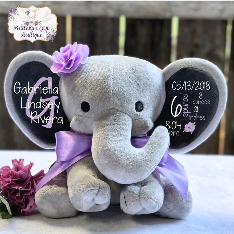 K&ouml;nnte beinhalten: Ein graues Pl&uuml;schelefant-Spielzeug mit einem violetten Satinband um den Hals und einer violetten Stoffblume auf dem Kopf. Das linke Ohr zeigt den Namen Gabriella Lindsey Rivera mit einem gro&szlig;en Buchstaben G, das rechte Ohr zeigt Geburtsdaten.