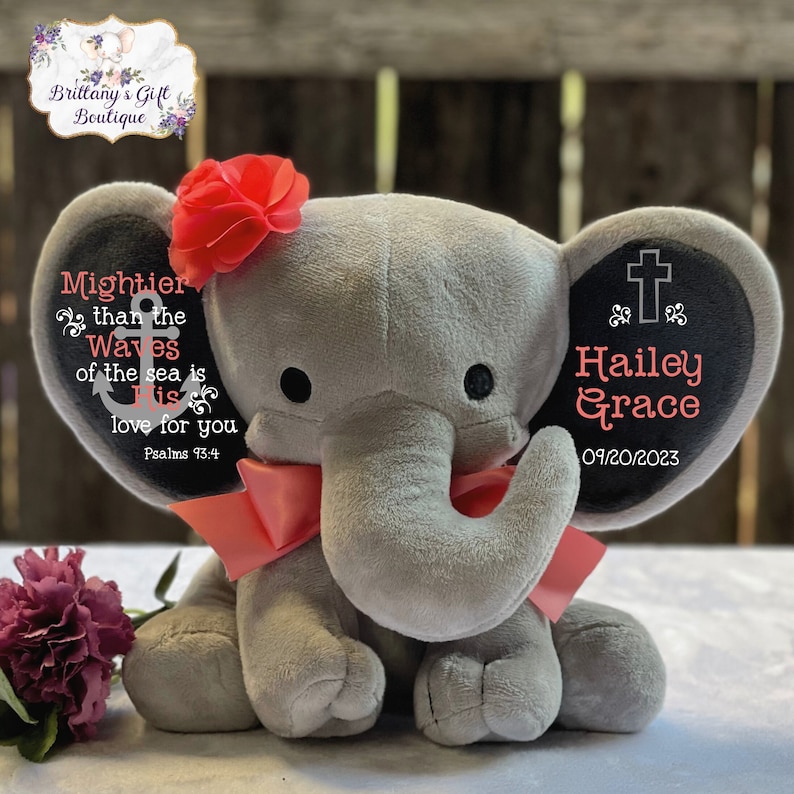 May include: A gray plush elephant featuring a coral-colored fabric flower on its head and a matching coral ribbon around its neck. The ears are personalized with black text reading "Mightier than the Waves of the sea is His love for you Psalms 93:4" and "Hailey Grace 09/20/2023" with a cross symbol.