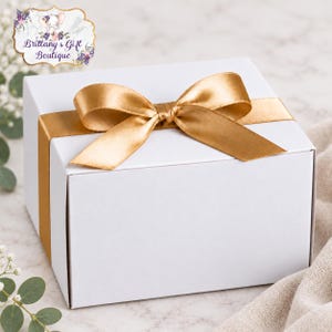 May include: A square white gift box wrapped with a smooth, metallic gold satin ribbon tied in a bow on top. The box features a clean, solid white exterior surface and is centered with the gold ribbon crossing horizontally across the lid.