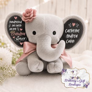 May include: A grey plush elephant toy with a pink satin ribbon around its neck and a pink fabric flower on its head. The left ear features the text Handpicked for earth by my Grandma in heaven, and the right ear displays the name Cathrine Andrea Cohn.