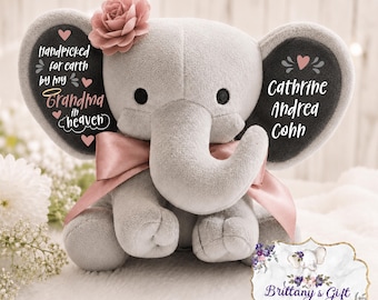 Puede incluir: Un juguete de peluche de elefante gris con una cinta de satén rosa alrededor del cuello y una flor de tela rosa en la cabeza. La oreja izquierda tiene el texto Handpicked for earth by my Grandma in heaven y la derecha el nombre Cathrine Andrea Cohn.