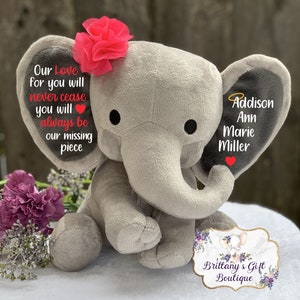 Może przedstawiać: Szary pluszowy słoń z r&oacute;żowym kwiatem na głowie. Słoń ma na jednym uchu napis "Our Love for you will never cease you will always be our missing piece." Drugie ucho ma napis "Addison Ann Marie Miller" z aureolą nad nazwiskiem. Słoń siedzi na białym tle z tabliczką z napisem "Brittany's Gift Boutique".