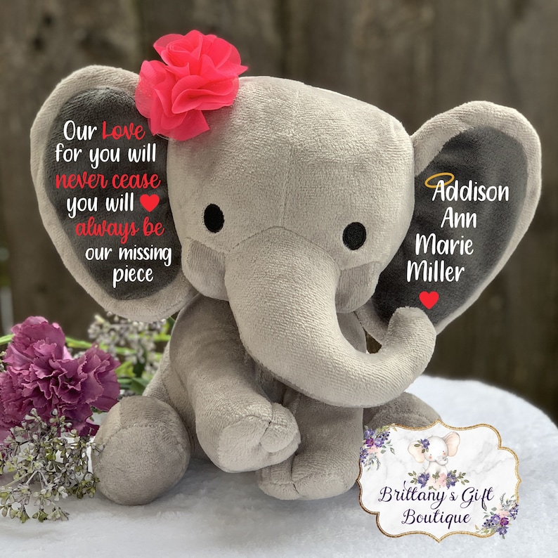 Może przedstawiać: Szary pluszowy słoń z r&oacute;żowym kwiatem na głowie. Słoń ma na jednym uchu napis "Our Love for you will never cease you will always be our missing piece." Drugie ucho ma napis "Addison Ann Marie Miller" z aureolą nad nazwiskiem. Słoń siedzi na białym tle z tabliczką z napisem "Brittany's Gift Boutique".