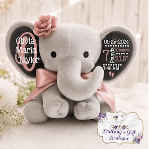 May include: A grey plush elephant toy featuring a pink satin bow and a pink fabric flower on its head. The ears display white text: Olivia Maria Taylor, 01-25-2024, 7 pounds, 10 ounces, 21.5 inches, and 7:48 AM, along with small pink footprint icons.