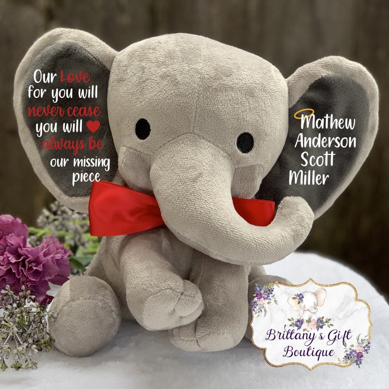 Może przedstawiać: Szary pluszowy słoń z czerwoną kokardką i czarnymi uszami. Słoń ma serce po lewej stronie i napis "Our Love for you will never cease you will always be our missing piece" na lewym uchu. Prawe ucho ma napis "Mathew Anderson Scott Miller". Słoń siedzi na białym tle z tabliczką z napisem "Brittany's Gift Boutique".