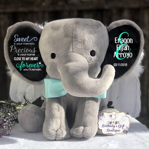 May include: A gray plush elephant with a light blue bow tie. The elephant's ears have text that reads "Sweet is your memory, Precious is your name, Close to my heart forever you'll remain." The other ear has the name "Eragon Elijah Arroyo" and the date "03/11/2019."