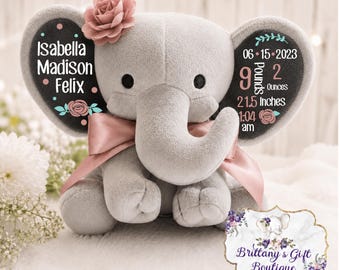 May include: A grey plush elephant toy with a pink satin bow and a pink fabric flower on its head. The ears feature black panels with white text: Isabella Madison Felix, 06-15-2023, 9 Pounds 2 Ounces, 21.5 Inches, and 1:04 am, accented with floral motifs.