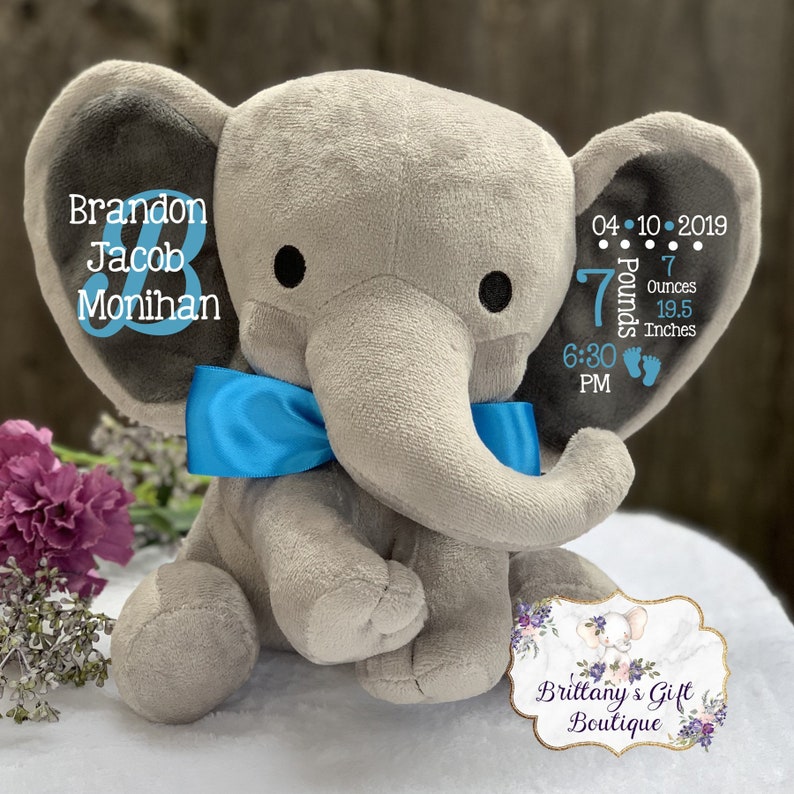 May include: A plush gray elephant toy with a bright blue satin bow around its neck. The elephant features personalized text including the name Brandon Jacob Monihan, the date 04-10-2019, 7 pounds 7 ounces, 19.5 inches, and 6:30 PM with blue accents.