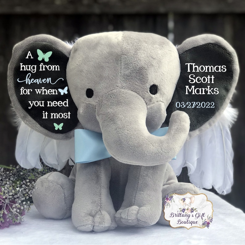 Puede incluir: Un elefante de peluche gris con alas de &aacute;ngel y una pajarita azul claro. Las orejas del elefante tienen el texto "A hug from heaven for when you need it most" y "Thomas Scott Marks 03/27/2022".