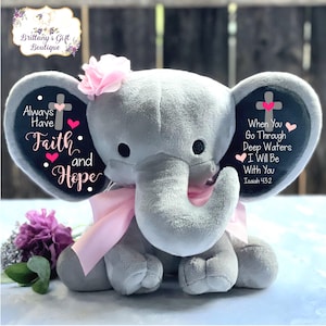 May include: A gray plush elephant with pink ears and a pink bow. The elephant has a black and white inscription on each ear. The left ear says "Always Have Faith and Hope" with a pink cross and hearts. The right ear says "When You Go Through Deep Waters I Will Be With You Isaiah 43:2" with a pink cross and hearts.