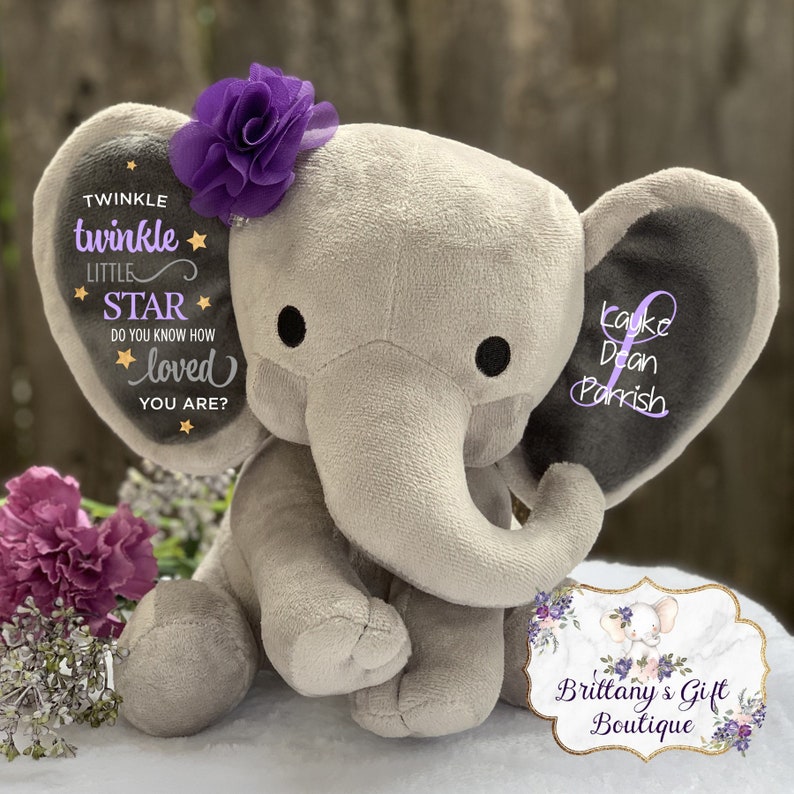 Puede incluir: Un elefante de peluche gris con una flor morada en la cabeza. Las orejas del elefante tienen el texto "Twinkle Twinkle Little Star" y "Kayke Dean Parrish". Un regalo para beb&eacute;s de Brittany's Gift Boutique.