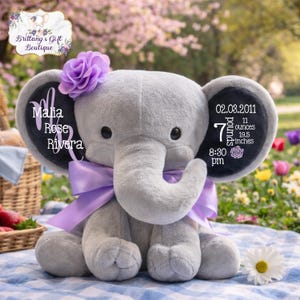 May include: A gray plush elephant with a purple bow, flower, and personalized text on the ears. The text includes a name, date, weight (7 pounds, 11 ounces), and length (19.5 inches). A charming keepsake.