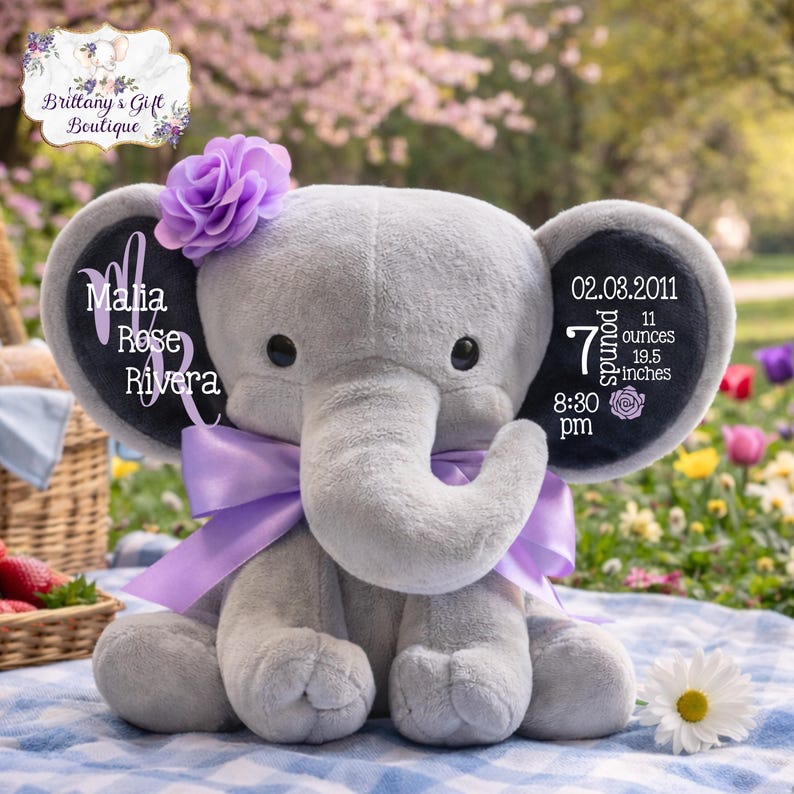 May include: A gray plush elephant with a purple bow, flower, and personalized text on the ears. The text includes a name, date, weight (7 pounds, 11 ounces), and length (19.5 inches). A charming keepsake.