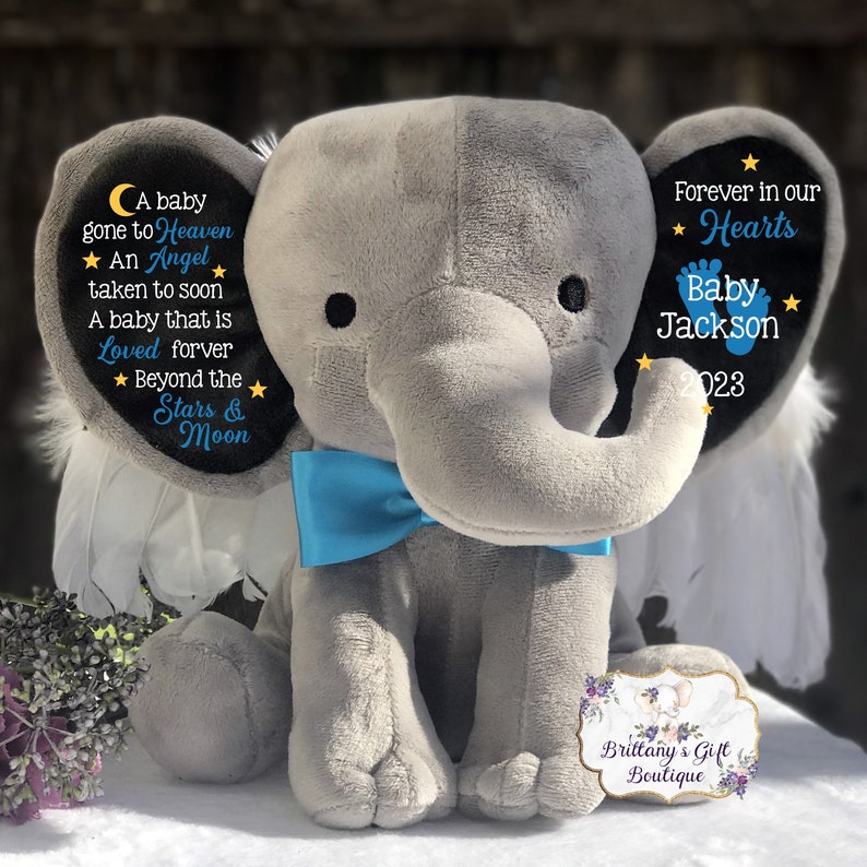 Puede incluir: Un elefante de peluche gris con una pajarita azul y alas blancas de plumas. El elefante tiene texto bordado en sus orejas: "Un beb&eacute; que se fue al cielo, Un &aacute;ngel que se fue demasiado pronto, Un beb&eacute; que es amado para siempre, M&aacute;s all&aacute; de las estrellas y la luna" y "Para siempre en nuestros corazones, Beb&eacute; Jackson 2023".