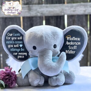 Może przedstawiać: Szary pluszowy słoń z niebieskimi skrzydłami i czarną filcową metką na ucho. Metka na ucho zawiera tekst "Our Love for you will never cease you will always be our missing piece" po jednej stronie i "Mathew Anderson Scott Miller" po drugiej.